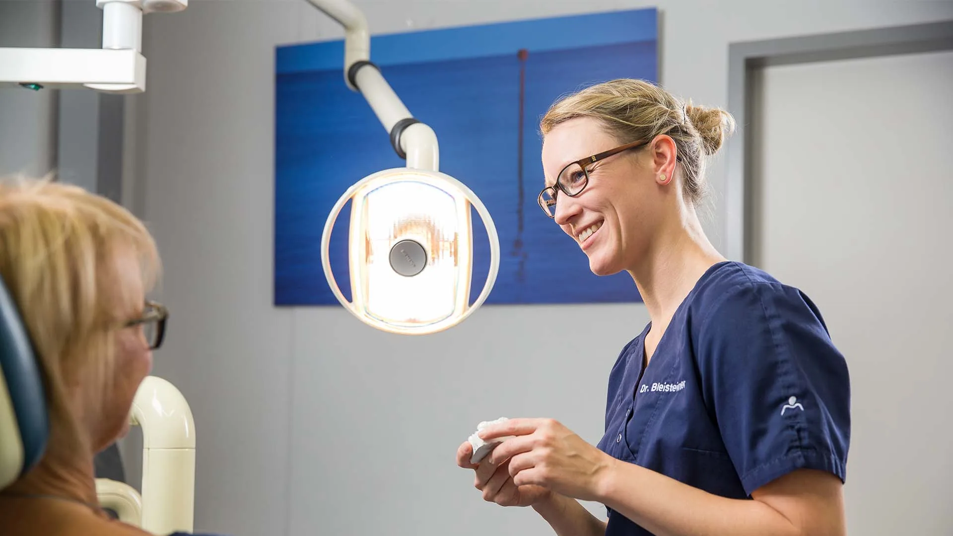 Zahnärztin in blauem Kittel mit Dr. Bleisteiner Schriftzug lächelt eine Patientin während der Behandlung an.