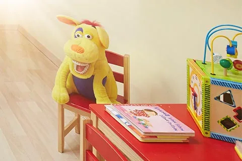 Plüschtier-Hund sitzt auf einem Kindersessel neben einem kleinen roten Tisch mit einem Stapel Kinderbücher und einem bunten Holzaktivitätsspielzeug.
